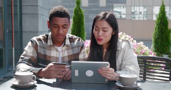 Happy Couple of African Man Holding a Credit Card and Cheerful Asian Woman with Tabletsitting at the alt