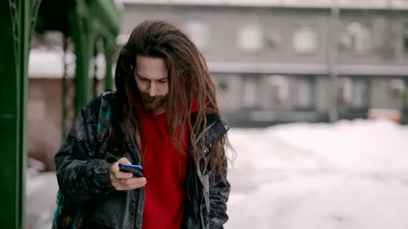 A View of a Bearded Man with Dreadlocks Looking at the Phone Screen and Laughing While Walking Down alt