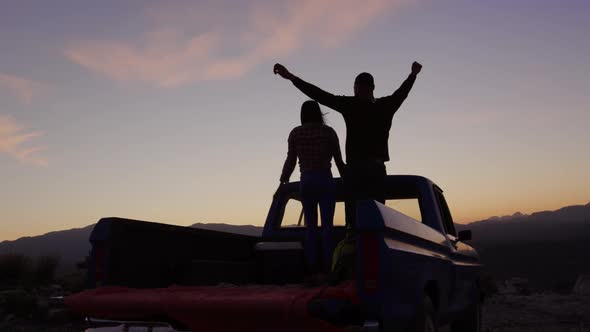 Young couple on a road trip sitting outside on their truck at dusk alt