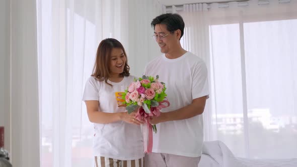 Asian man surprising his lover by giving a flower bouquet in bedroom. alt