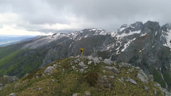 A Hiker Stands on Top of a Mountain and Looks at Snow-capped Mountains, Raising His Hands in Victory