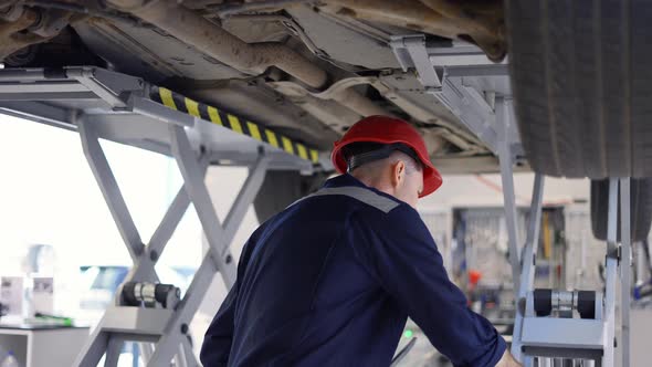A Car Mechanic in Helmet Standing Under Lifted Car Making Notes on a Tablet alt