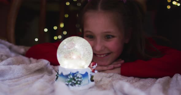 Caucasian girl smiling and looking at snow globe while lying under blanket fort during christmas at alt