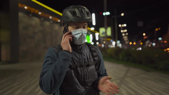 Cyclist Delivery Man Wearing Medical Mask and Helmet Speaks to Next Customer on Phone Before Leaving alt