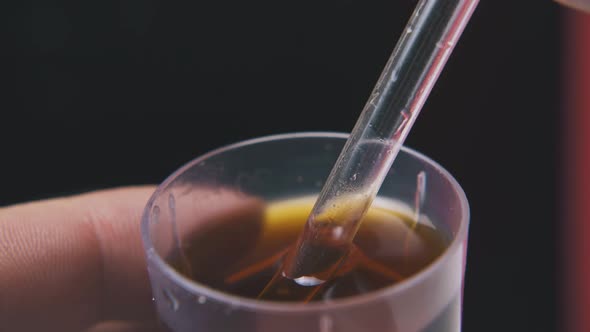 Person Puts Pipette Into Brown Liquid on Dark Background alt