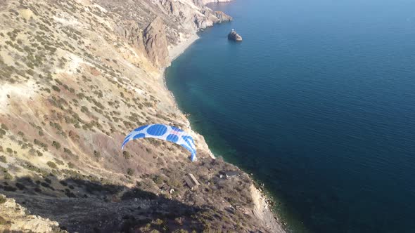 Aerial Drone View of a Man Flying a White and Blue Paraglider Over a Hill and Trees to the Sea Waves alt
