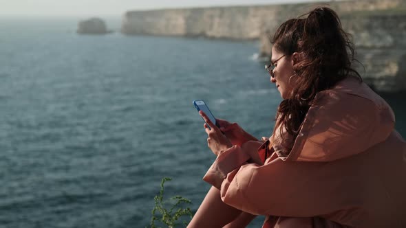 Woman is Communicating By Online Chat in Smartphone Sitting on High Sea Shore alt