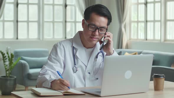 Asian Male Doctor Talking On Phone And Writing On Notebook At Home Office alt