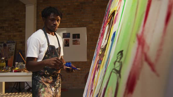 African american male artist wearing apron painting with palette knife on canvas at art studio alt