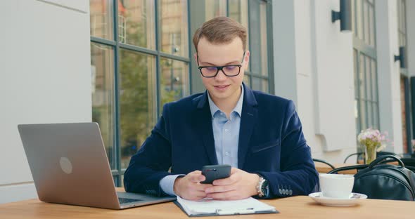 A Businessman Is Sitting at a Table in a Cafe and Texting on His Smartphone alt