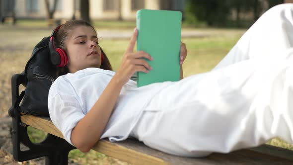 Relaxed Positive Young Woman Lying on Bench at College Yard Listening Music in Headphones Singing alt