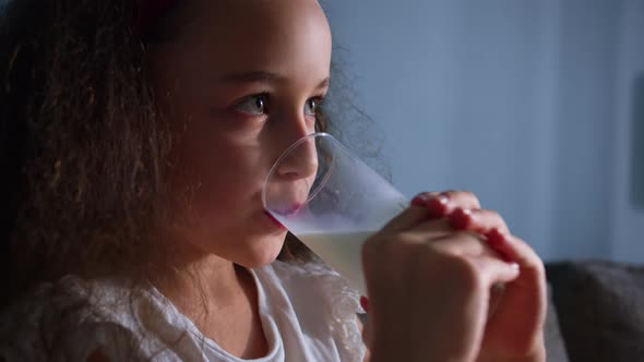 Little Girl Positive Child Drinks Milk While Sitting on Sofa at Home While Watching TV alt