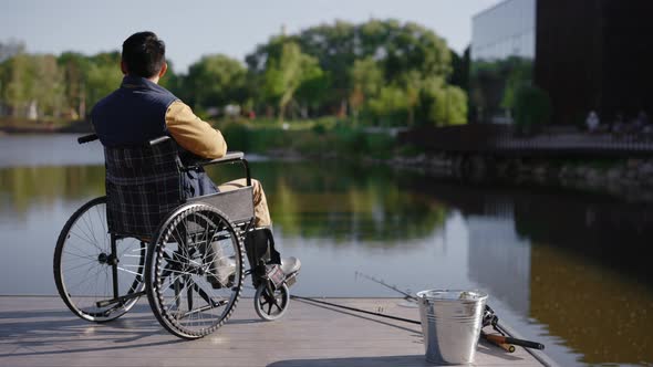 Back View of the Disabled Fisherman Sitting on the Wheelchair in the Pier Looking at the Local Lake alt