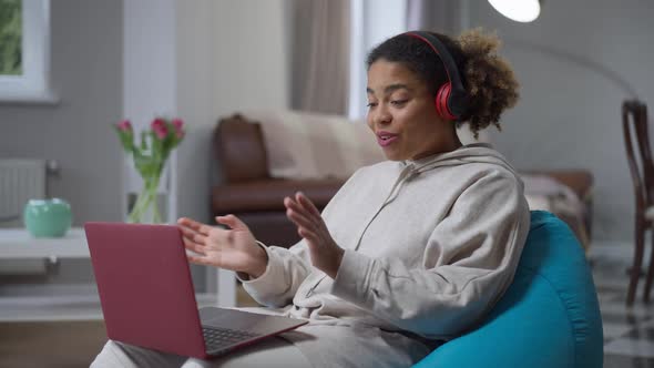 Side View Portrait of Positive Young African American Woman in Headphones Waving at Laptop Webcam alt