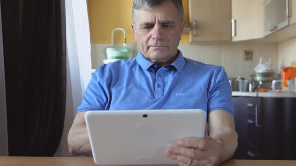 Aged Male At The Blue Shirt Sits And Uses A White Tablet Pc At Home