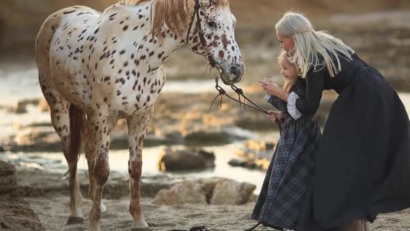 Woman in Classic Dress Standing on Coast and Bonding with Little Daughter and Mottled Horse