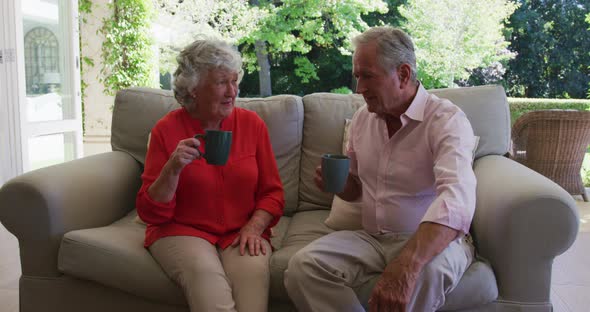 Happy caucasian senior couple on couch in sunny living room talking and drinking cups of coffee alt