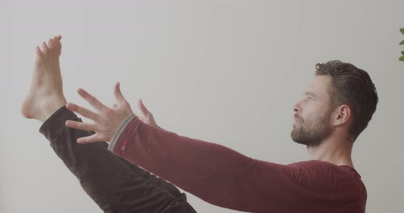 Side Portrait of Bearded Man Balancing in Seated Asana Pose Indoors on White Wall Background alt