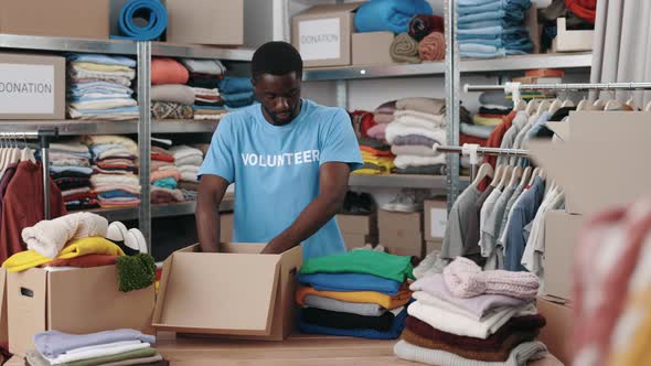 African American Man Standing at the Warehouse While Sorting and Iterating Clothes for Donations alt