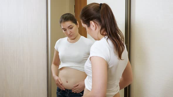 Young Overweight Woman in Front of a Mirror Palpates Her Sides and Stomach alt