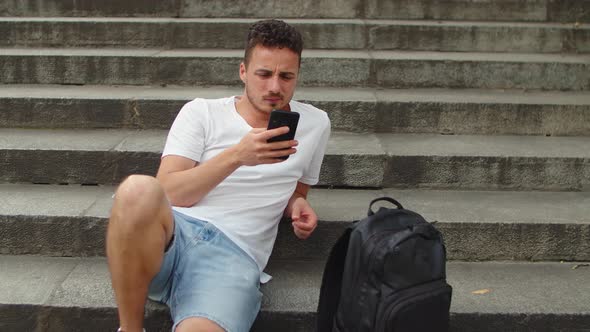Handsome Young Man Sitting on the Steps of the Stairs in the City with Smartphone in Hand alt