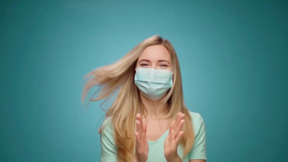 Woman in Medical Mask on Face is Very Happy and Claps Her Hands Against Blue Background alt