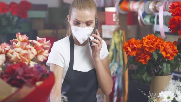 Florist Woman in a Protective Medical Mask Discusses the Order By Talking on the Smartphone alt