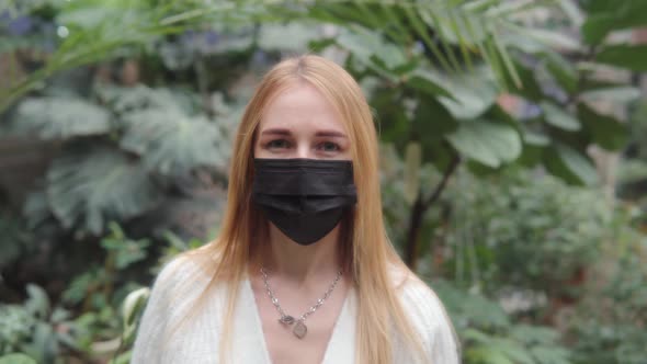 Portrait of Young Woman in Medical Mask Standing Against the Background of Plants in the Greenhouse