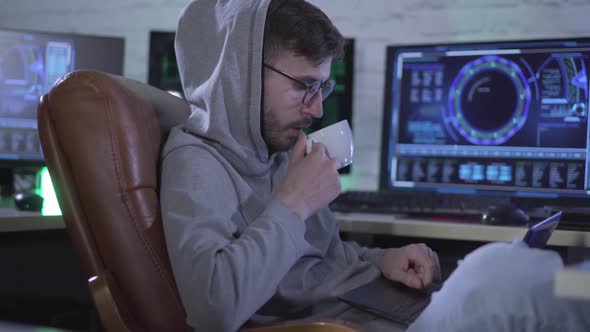 Side View of Confident Caucasian Programmer Typing on Laptop Keyboard and Drinking Coffee at Night alt