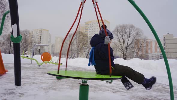 A Little Boy in the Winter Spins on a Swing on the Playground