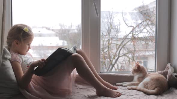 The Baby Girl Reading a Book Sitting on the Windowsill alt