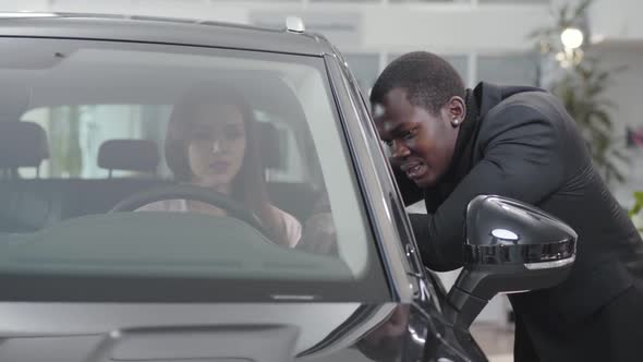Portrait of Smiling African American Car Dealer Talking To Young Caucasian Woman Sitting in Car alt