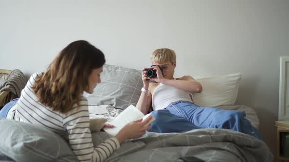 Young Lesbian Couple Takes Pictures and Reads While Lying on Bed in Bedroom alt