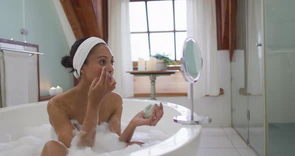Mixed race woman taking a bath applying beauty face mask alt