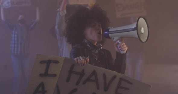 Diverse group of people wearing face masks holding protest signs shouting into megaphone alt