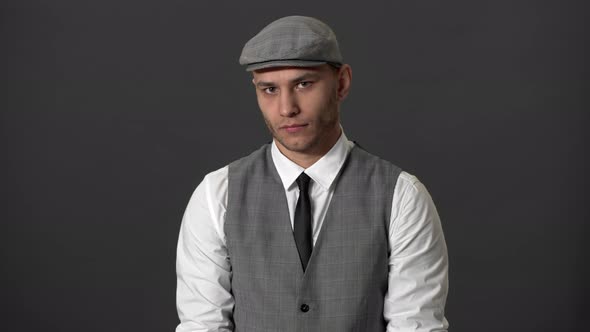 Portrait of Stylish Guy Wearing Suit and Flat Cap Posing on Camera with Meaningful Look Over Grey alt