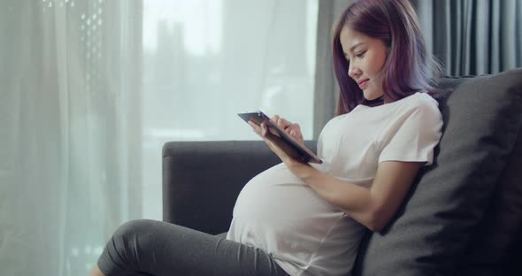 Beautiful Asian pregnant woman stroking belly while sitting on a sofa in the living room at home.