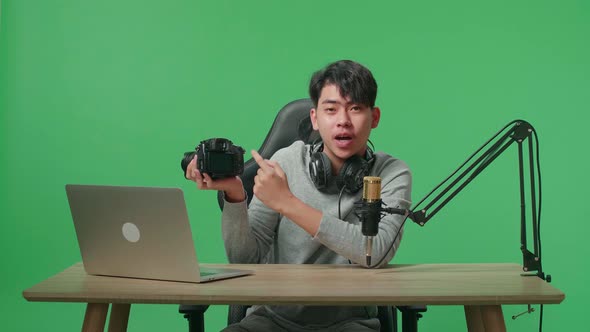 Asian Man With Computer Reviewing Camera While Sitting In Front Of Green Screen Background alt