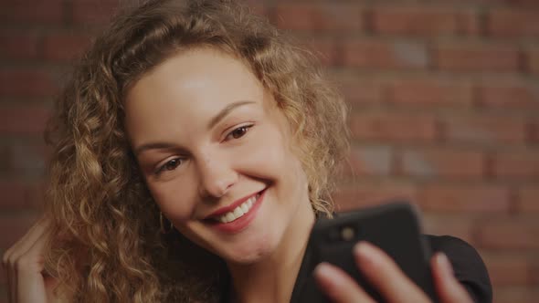 Portrait of a Young Curly Caucasian Business Woman Taking Selfies and Smiling with a Red Brick Wall alt