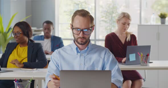 Young Diverse Office Managers or Designers Sitting at Desks and Using Laptop and Smiling at Camera alt