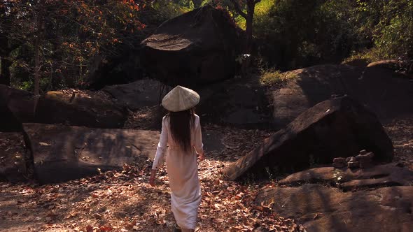 Young woman in white dress and vietnamese hat walking in forest near big stones Vat Phou Temple Laos alt