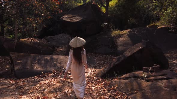 Young woman in white dress and vietnamese hat walking in forest near big stones Vat Phou Temple Laos