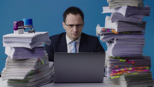 Serious Manager Working on Laptop Among Piles of Paperwork, Overloaded with Work alt