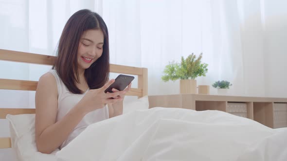 Young Asian woman using smartphone checking social media feeling happy smiling while lying on bed. alt