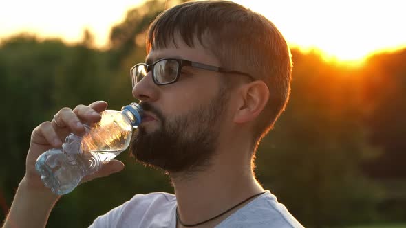 Man Holds Bottle of Water and Drinks Water on Background of Green Summer Park at Sunset
