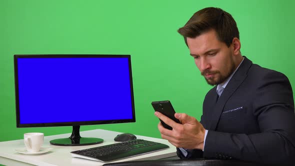 A Young Businessman Sits in Front of a Computer and Works on a Smartphone - Green Screen Studio alt