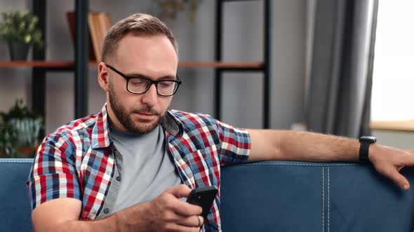 Pensive Modern Guy Typing Message Browsing Internet Net Web Site Connection Use Smartphone on Sofa alt