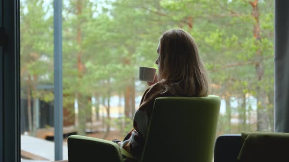 woman drinking tea sitting in a chair by the window with a view of the forest