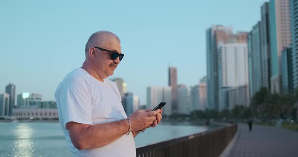 Bearded Senior Man Standing Outdoors Using Smartphone. Retired Male Reading News, Chatting with His alt