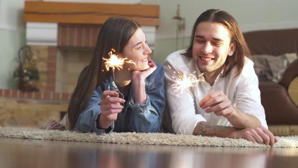 Joyful Young Beautiful Guy and Girl with Sparklers in Hands Lying on the Carpet alt